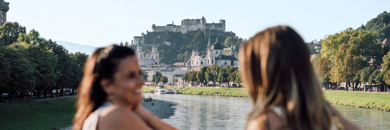 Two friends on Müllnersteg in Salzburg, with Hohensalzburg Fortress in the background. | © SalzburgerLand Tourismus, Chris Perkles
