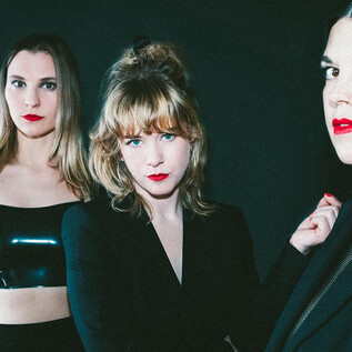 Three women pose in elegant clothing against a dark background. They have a strong gaze and wear bold lipstick. | © Maren Michaelis