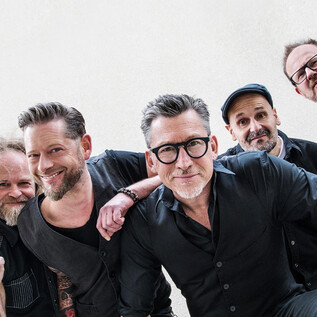 A band is happily posing for a photo. It consists of six members who are wearing casual clothing and are looking friendly at the camera. | © Franz Schepers