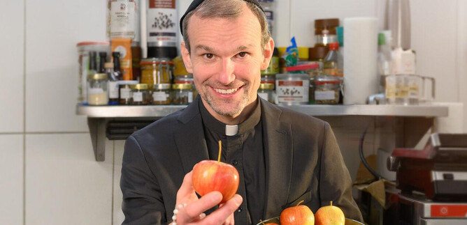 A smiling man in a black outfit is holding an apple and standing in front of a modern kitchen setting. In the background, various foods and kitchen utensils can be seen. | © SLT / Christian Krautzberger