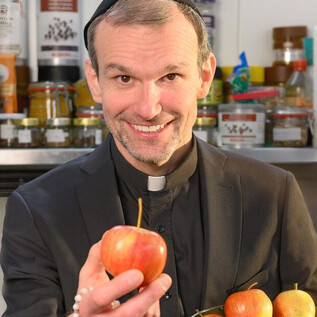 A smiling man in a black outfit is holding an apple and standing in front of a modern kitchen setting. In the background, various foods and kitchen utensils can be seen. | © SLT / Christian Krautzberger