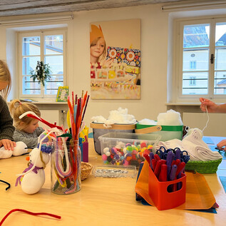 Two children are crafting with colorful materials at the table, while an adult helps them. The room is bright and cheerful, with many crafting supplies scattered about. | © Altstadt Salzburg
