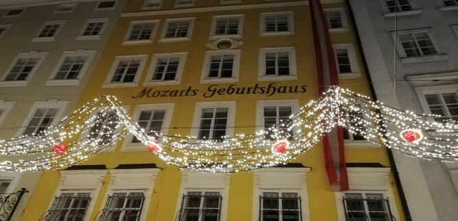 The image shows Mozart's birthplace, illuminated with festive decoration. The facade is yellow and contrasts with the night. | © ISM
