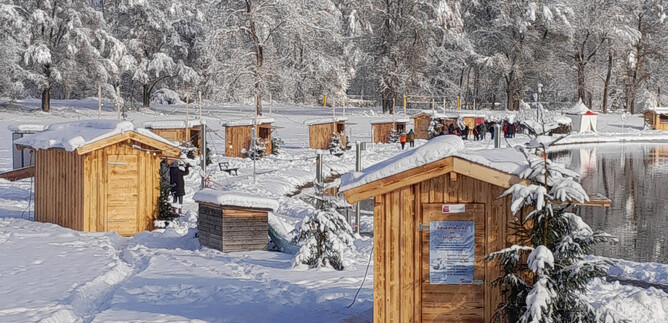 A snowy landscape with small wooden cabins around a frozen lake. The sky is clear and the trees are covered with snow. | © Adventmarkt am Salzachsee