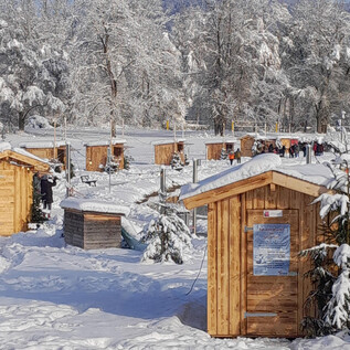 A snowy landscape with small wooden cabins around a frozen lake. The sky is clear and the trees are covered with snow. | © Adventmarkt am Salzachsee