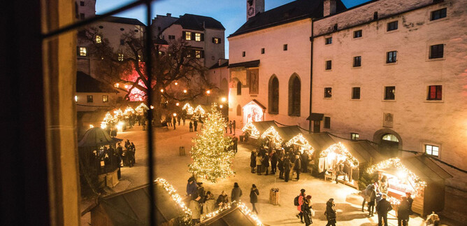 A festive Christmas market with a decorated tree and illuminated stalls. Many people enjoy the atmosphere on a clear evening. | © Burgen und Schlösser
