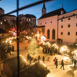 A festive Christmas market with a decorated tree and illuminated stalls. Many people enjoy the atmosphere on a clear evening. | © Burgen und Schlösser