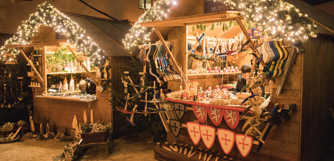 Ein festlich geschmückter Weihnachtsmarkt mit Holzständen. An einem Stand werden Spielzeuge und Souvenirs angeboten, während Lichter für eine gemütliche Atmosphäre sorgen.