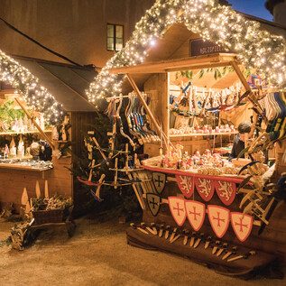 Ein festlich geschmückter Weihnachtsmarkt mit Holzständen. An einem Stand werden Spielzeuge und Souvenirs angeboten, während Lichter für eine gemütliche Atmosphäre sorgen.