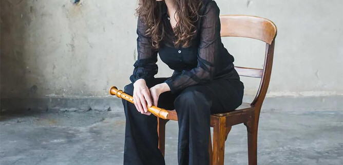 A woman with long hair is sitting on a wooden chair and smiling. She is wearing a black top and black pants, while holding a flute. | © Henning Ross