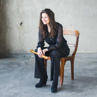 A woman with long hair is sitting on a wooden chair and smiling. She is wearing a black top and black pants, while holding a flute. | © Henning Ross