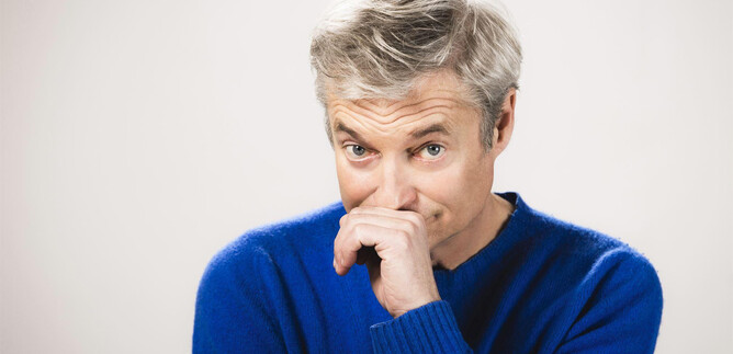 A man with gray hair is wearing a blue sweater. He has a thoughtful expression and is touching his face. | © Moritz Schell
