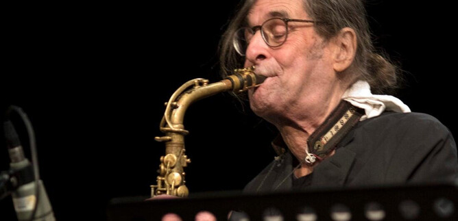 An older musician is playing the saxophone on a stage. He is wearing glasses and has long hair. | © J.P. Sonntag