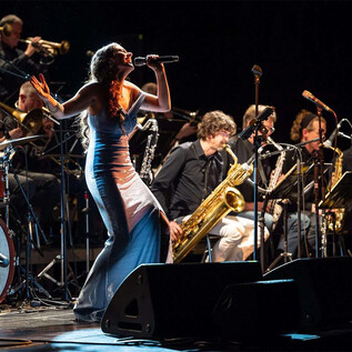 A singer in an elegant dress performs on stage in front of a band. In the background, musicians play various wind instruments. | © Albert Moser