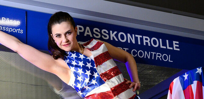 A woman in a dress designed with the American flag poses in front of a passport control area. The background features the "Passport Control" sign in several languages. | © SLT / Tobias Witzgall