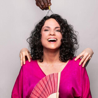 A woman with curly hair is wearing a bright pink top and holding a fan. In the background, two hands can be seen gently supporting her. | © Jasmin Schuller