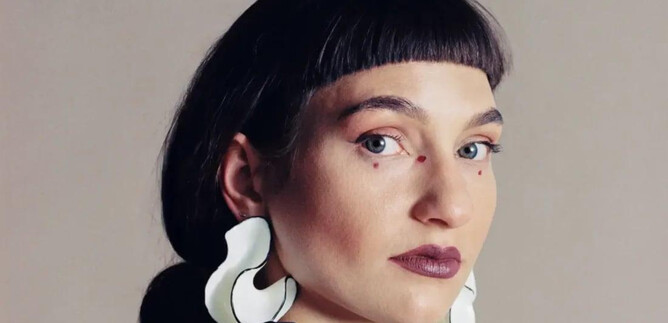 A person with short, dark hair and striking white earrings looks directly at the camera. The background is neutral and highlights the focus on the face. | © Anna Hadaier