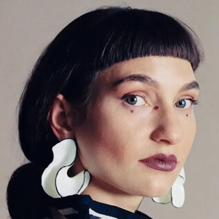 A person with short, dark hair and striking white earrings looks directly at the camera. The background is neutral and highlights the focus on the face. | © Anna Hadaier