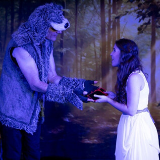 A man in a bear costume hands a woman in a white dress a red object. The scene takes place in a mystical forest. | © Theater mit Seele