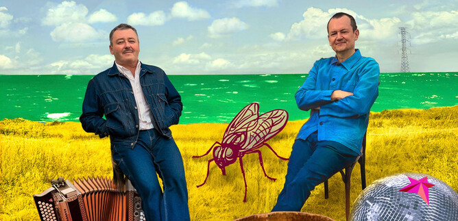 Two men are sitting in nature, surrounded by a colorful landscape. In the background, you can see the sea and a large fly made of art. | © Helmut Wimmer