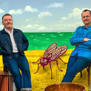 Two men are sitting in nature, surrounded by a colorful landscape. In the background, you can see the sea and a large fly made of art. | © Helmut Wimmer
