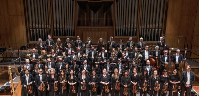 A large orchestra is standing on stage in formal attire. In the background, tall organ pipes can be seen. | © recreation Event GmbH