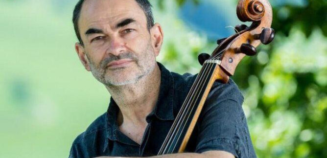 A man with a cello sits outdoors in front of a green landscape. He is wearing a black shirt and is looking kindly at the camera. | © Stefano Veggetti