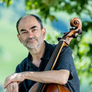 A man with a cello sits outdoors in front of a green landscape. He is wearing a black shirt and is looking kindly at the camera. | © Stefano Veggetti