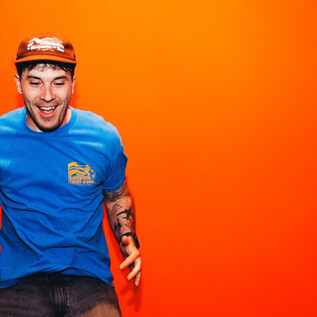A young man wearing a blue T-shirt and a red cap is standing in front of an orange wall and smiling. He looks cheerful and energetic. | © Rockhouse Salzburg