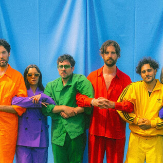 Sechs Personen stehen vor einem blauen Hintergrund und tragen bunte Outfits in Gelb, Grün, Orange und Lila. Sie posieren zusammen und scheinen eine fröhliche Atmosphäre auszustrahlen. | © Paul Vincent Schütz