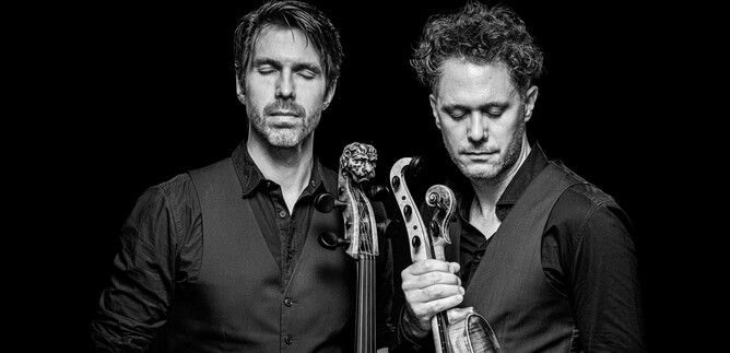 Two musicians stand close together, with serious expressions. They hold musical instruments in their hands and are depicted against a dark background. | © Stephan Doleschal