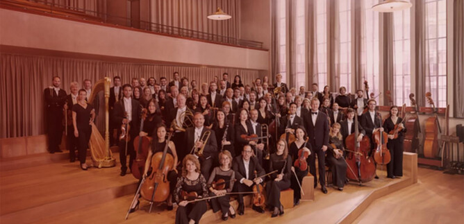 A large orchestra is standing in a room with high windows. The musicians are wearing formal attire and holding their instruments. | © Noë_Flum@