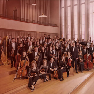 A large orchestra is standing in a room with high windows. The musicians are wearing formal attire and holding their instruments. | © Noë_Flum@