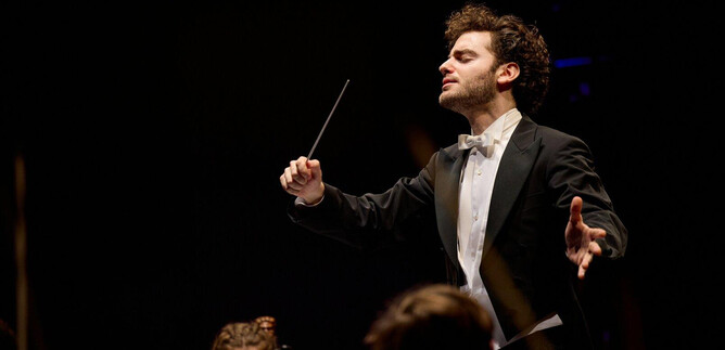 A conducting musician in an elegant suit leads an orchestra. The atmosphere is festive and full of musical energy. | © Oliver Borchert