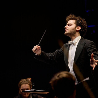 A conducting musician in an elegant suit leads an orchestra. The atmosphere is festive and full of musical energy. | © Oliver Borchert