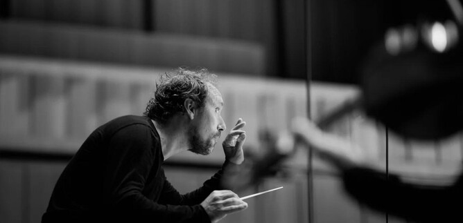 A conductor stands at the center and gestures passionately. In the background, musicians can be seen responding to his instructions. | © Valerie Spanjers