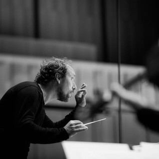 A conductor stands at the center and gestures passionately. In the background, musicians can be seen responding to his instructions. | © Valerie Spanjers