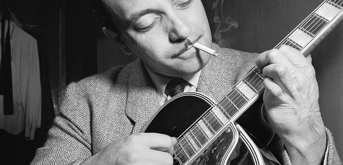 A man plays guitar while smoking a cigarette. He is wearing a suit and has a focused expression. | © William P. Gottlieb | Adam Cuerden
