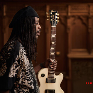Ein Musiker steht mit einer Gitarre in der Hand. Er trägt ein auffälliges Hemd und blickt nachdenklich in die Ferne. | © Tino Sieland