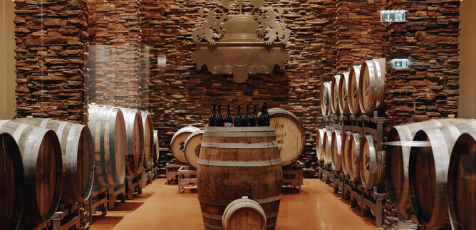 A wine cellar with wooden barrels and a central barrel. The walls are lined with stones, creating a warm atmosphere. | © Sarah Matzer Photography / www.designmatzer.at