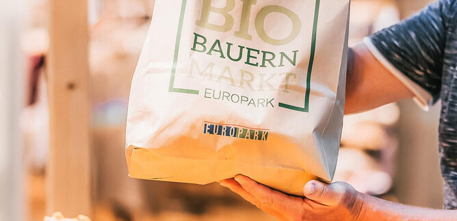 A person is holding a shopping bag with the inscription "ORGANIC FARMERS MARKET EUROPE". The bag is made of eco-friendly material and contains fresh, organic products. | © LOOP