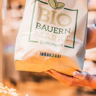 A person is holding a shopping bag with the inscription "ORGANIC FARMERS MARKET EUROPE". The bag is made of eco-friendly material and contains fresh, organic products. | © LOOP