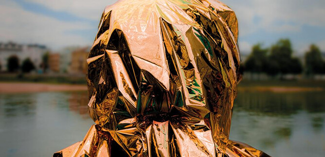 A person stands by the water wearing a shiny, golden coat. The sky is cloudy, and it is a calm environment. | © Valentina Schuster