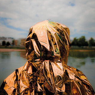 A person stands by the water wearing a shiny, golden coat. The sky is cloudy, and it is a calm environment. | © Valentina Schuster