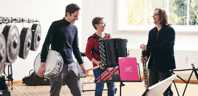 Three musicians stand in a bright room and play different instruments. One plays the accordion, while the others hold a drum and a woodwind instrument. | © Andreas Hechenberger
