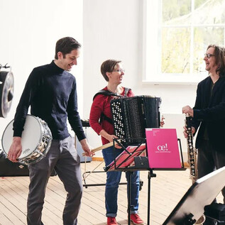 Three musicians stand in a bright room and play different instruments. One plays the accordion, while the others hold a drum and a woodwind instrument. | © Andreas Hechenberger