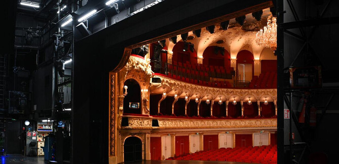 A glimpse behind the scenes of a theater with red seats and an elegant decor. The stage is illuminated and showcases magnificent details. | © Neumayr_Leo