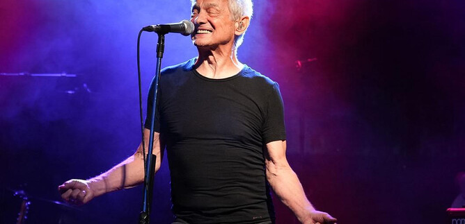 An older man stands on stage and sings into a microphone. Colorful lights and fog create a vibrant concert atmosphere. | © Paul Trondl