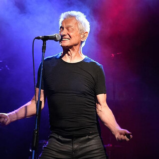An older man stands on stage and sings into a microphone. Colorful lights and fog create a vibrant concert atmosphere. | © Paul Trondl