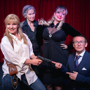 A group of four actors stands on a stage. They pose in different characters, a woman holding a gun in her hand. | © Theater Dinner Company